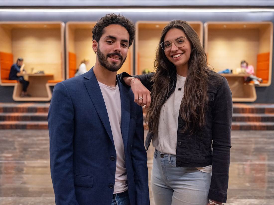 Joven con saco azul y camiseta clara posando junto a una joven de cabello largo y gafas, vestida con chaqueta negra y camiseta blanca, ambos sonriendo frente a un moderno espacio universitario con cubículos iluminados al fondo donde otros estudiantes estudian o conversan.